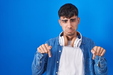 Young hispanic man standing over blue background pointing down looking sad and upset, indicating...
