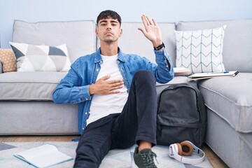 Young hispanic man sitting on the floor studying for university swearing with hand on chest and...