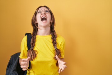 Young caucasian woman wearing student backpack over yellow background angry and mad screaming frustrated and furious, shouting with anger. rage and aggressive concept. - obrazy, fototapety, plakaty
