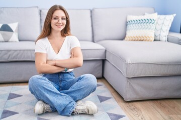 Young caucasian woman sitting on the floor at the living room happy face smiling with crossed arms looking at the camera. positive person.