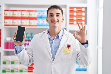 Handsome hispanic man working at pharmacy drugstore showing smartphone screen doing ok sign with fingers, smiling friendly gesturing excellent symbol