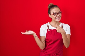 Young hispanic woman wearing waitress apron over red background amazed and smiling to the camera...