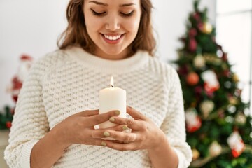 Young beautiful hispanic woman wearing holding candle standing by christmas tree at home