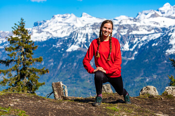 Naklejka premium sweet woman with pigtails stretches and does yoga on top of a mountain in the swiss alps enjoying the view of the snow-capped peaks; yoga position dancer; spring hike in the swiss alps, interlaken