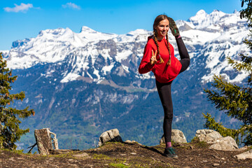 Naklejka premium sweet woman with pigtails stretches and does yoga on top of a mountain in the swiss alps enjoying the view of the snow-capped peaks; yoga position dancer; spring hike in the swiss alps, interlaken