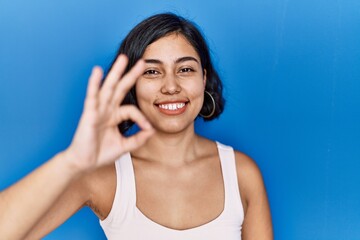 Fototapeta premium Young hispanic woman standing over blue background smiling positive doing ok sign with hand and fingers. successful expression.