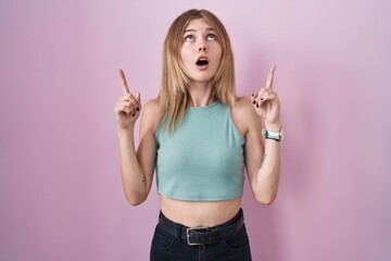 Blonde caucasian woman standing over pink background amazed and surprised looking up and pointing with fingers and raised arms.