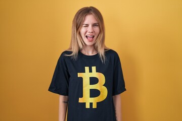 Blonde caucasian woman wearing bitcoin t shirt winking looking at the camera with sexy expression,...