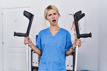 Young physiotherapist woman holding crutches in shock face, looking skeptical and sarcastic, surprised with open mouth