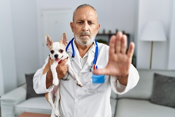 Mature veterinarian man checking dog health with open hand doing stop sign with serious and...