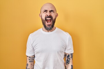 Young hispanic man with tattoos standing over yellow background angry and mad screaming frustrated...