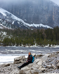 A hiker girl sits on rocks at a famous mountain lake in switzerland during gloomy weather with cloudy mountain peaks in the background; fog at Lake Oeschinensee in the swiss alps