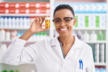 African american woman pharmacist smiling confident holding pills bottle at pharmacy