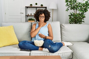 African american woman with afro hair sitting on the sofa eating popcorn at home covering mouth with hand, shocked and afraid for mistake. surprised expression