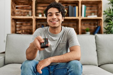 Young hispanic man watching tv sitting on the sofa at home.