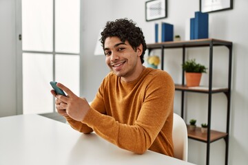 Young hispanic man using smartphone sitting on the table at home.