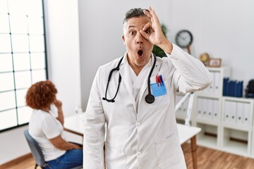 Middle age doctor man at the clinic with a patient doing ok gesture shocked with surprised face, eye looking through fingers. unbelieving expression.