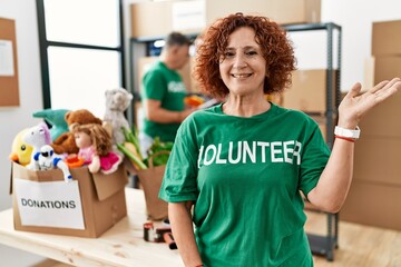 Middle age woman wearing volunteer t shirt at donations stand smiling cheerful presenting and pointing with palm of hand looking at the camera.