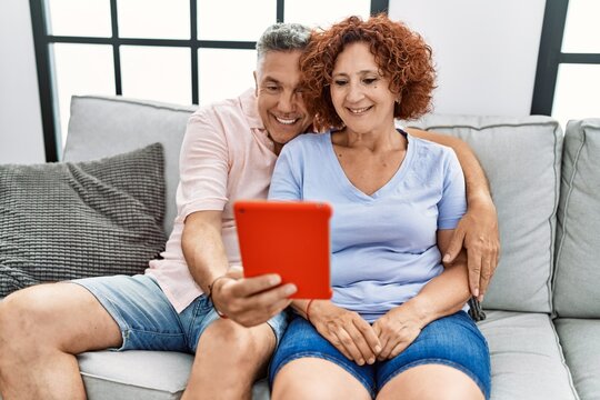 Middle age man and woman couple using touchpad hugging each other sitting on sofa at home