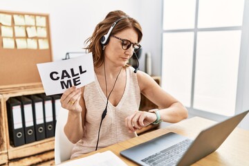 Middle age brunette woman wearing operator headset holding call me banner checking the time on...