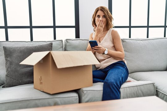 Middle age woman sitting on the sofa checking delivery package on the phone covering mouth with hand, shocked and afraid for mistake. surprised expression