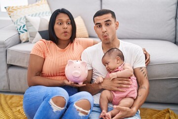 Young hispanic couple with baby holding piggy bank making fish face with mouth and squinting eyes, crazy and comical.