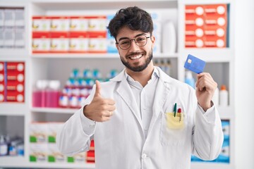 Hispanic man with beard working at pharmacy drugstore holding credit card smiling happy and positive, thumb up doing excellent and approval sign