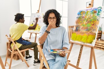 Young hispanic woman at art studio smelling something stinky and disgusting, intolerable smell,...