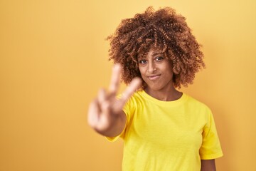 Young hispanic woman with curly hair standing over yellow background smiling looking to the camera showing fingers doing victory sign. number two.