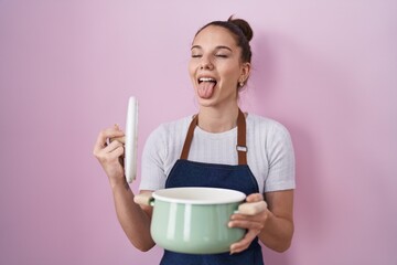 Young hispanic girl wearing apron holding cooking pot sticking tongue out happy with funny expression.