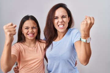 Young mother and daughter standing over white background angry and mad raising fist frustrated and furious while shouting with anger. rage and aggressive concept.