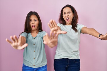 Young mother and daughter standing over pink background doing stop gesture with hands palms, angry and frustration expression