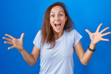 Brunette woman standing over blue background celebrating crazy and amazed for success with arms raised and open eyes screaming excited. winner concept