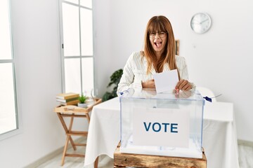 Hispanic business woman voting putting envelop in ballot box sticking tongue out happy with funny...