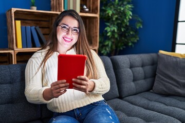 Young beautiful hispanic woman using touchpad sitting on sofa at home