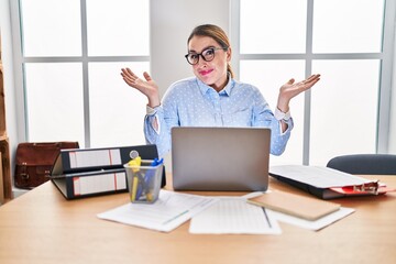 Young hispanic woman working at the office wearing glasses clueless and confused expression with...