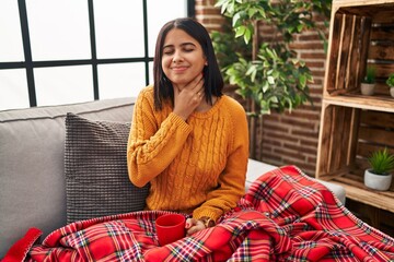 Young hispanic woman sitting on the sofa drinking a coffee at home touching painful neck, sore throat for flu, clod and infection