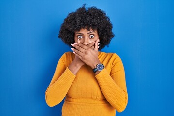 Fototapeta na wymiar Black woman with curly hair standing over blue background shocked covering mouth with hands for mistake. secret concept.
