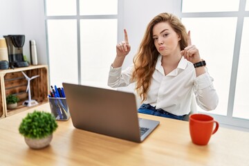 Young caucasian woman working at the office using computer laptop pointing up looking sad and...