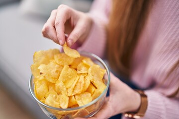 Young caucasian woman eating chips potatoes sitting on sofa at home