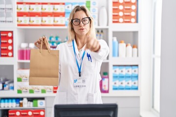 Young blonde woman working at pharmacy drugstore holding paper bag pointing with finger to the camera and to you, confident gesture looking serious