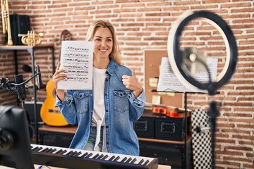 Young caucasian woman doing online music tutorial showing music sheet smiling happy and positive, thumb up doing excellent and approval sign