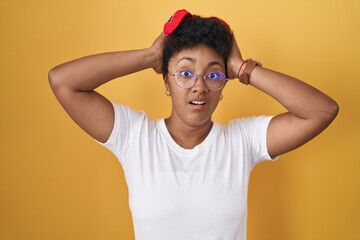 Young african american woman standing over yellow background crazy and scared with hands on head, afraid and surprised of shock with open mouth