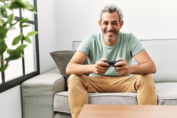 Fototapeta premium Middle age hispanic man playing video game sitting on the sofa looking positive and happy standing and smiling with a confident smile showing teeth