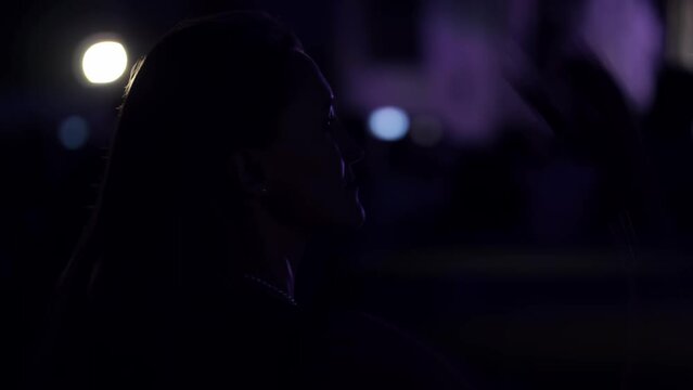 Woman is glancing at big scene of open air concert of an rock band and then she starting dancing. Singers perfomance outdoors. Stage is full of special effects and different colourful luminescences