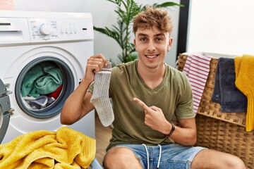 Young caucasian man doing laundry holding sock smiling happy pointing with hand and finger
