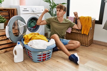 Young caucasian man putting dirty laundry into washing machine showing arms muscles smiling proud....