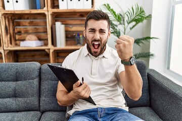 Handsome hispanic man holding clipboard working at psychology clinic angry and mad raising fist...