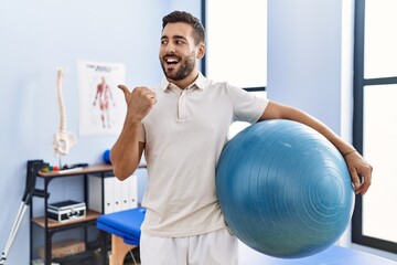Handsome hispanic man holding pilates ball at rehabilitation clinic pointing thumb up to the side...