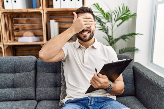Handsome hispanic man holding clipboard working at psychology clinic smiling and laughing with hand on face covering eyes for surprise. blind concept.